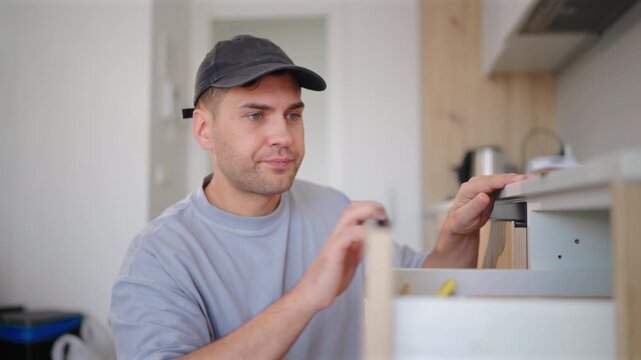 Skilled worker with toolbox performs maintenance in kitchen of home for repair project. Man wearing cap uses screwdriver to assemble wooden furniture with focused concentration and precision.