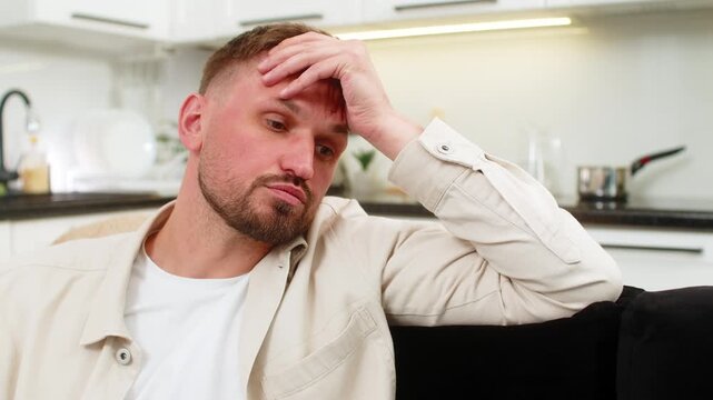 Young man at home stares ahead with tired mood and long sigh after many issues and missed goals. Caucasian guy on sofa rests hands on knees and shows sadness stress and dull vibe before short pause