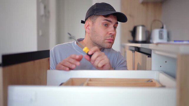 Professional worker with toolbox performs maintenance and repair work in kitchen of home. Man wearing cap uses screwdriver to assemble wooden cabinets and takes notes on clipboard in modern interior.