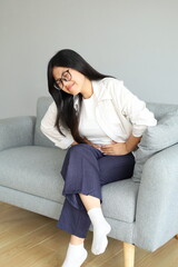 Obraz premium A young woman sits on the living room sofa, holding her stomach with a pained expression, feeling menstrual pain or stomach cramps.