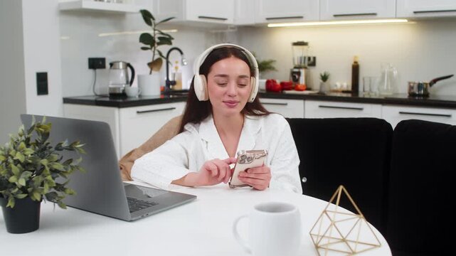 Young woman at home wears wireless headphones types message on smartphone takes break from tasks. Caucasian girl on sofa sits at table with laptop checks social app replies to chat relaxes happy mood.