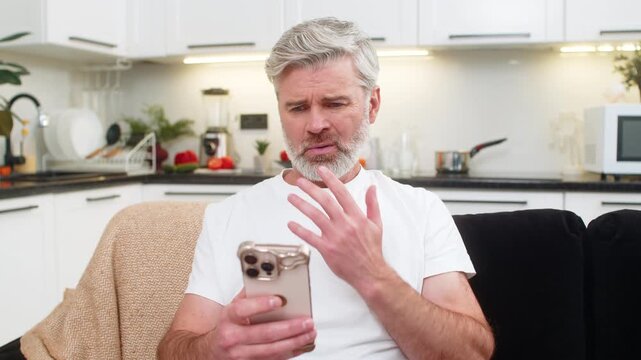 Middle-aged man at home on smartphone sees failed play game or job denial news sighs and shakes head no. Mature guy on sofa lowers gaze breath heavy pauses and tries to calm emotion and accept result