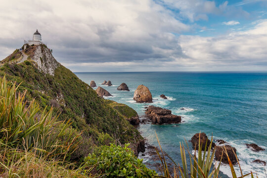 View of Nugget Point Lighthouse on a steep cliff overlooking rocky islets and the turquoise ocean under a cloudy sky in Ahuriri Flat, Otago Region, New Zealand.