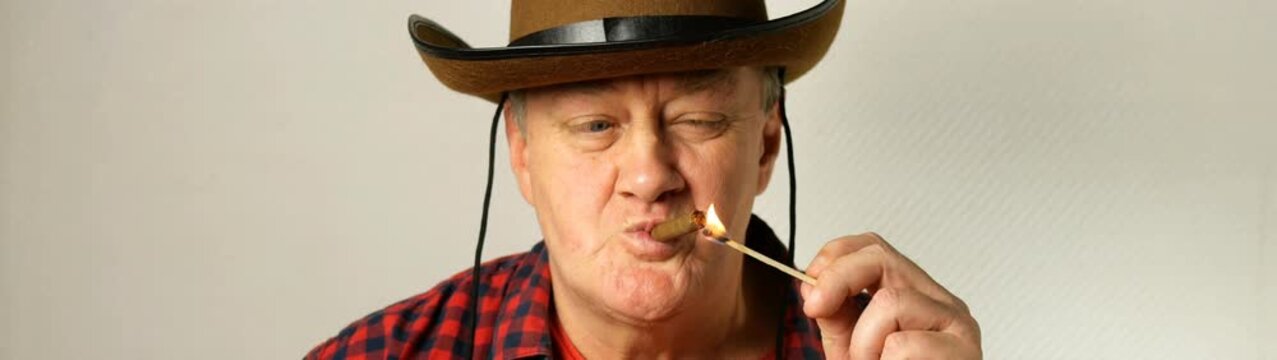 Mature man wearing a cowboy hat and plaid shirt lighting a cigar with a matchstick and creating smoke.