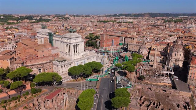 Cinematic aerial view in Rome Italy showcasing the majestic white marble Altare della Patria monument, Piazza Venezia, ancient Roman ruins, and terracotta rooftops under a clear blue summer sky.