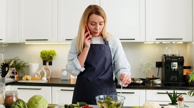 Middle-aged woman talks on smartphone while mixing vegetable salad for keto diet in home kitchen. Young girl shares simple healthy recipe with friend on call and keeps focused smile during lunch prep.