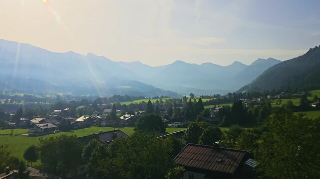 Beautiful cable car ride over Kitzbuhel, Austria, with alpine homes, rolling meadows, dark forested hills, and hazy mountain ranges in warm golden sunlight for travel footage in the Alps.