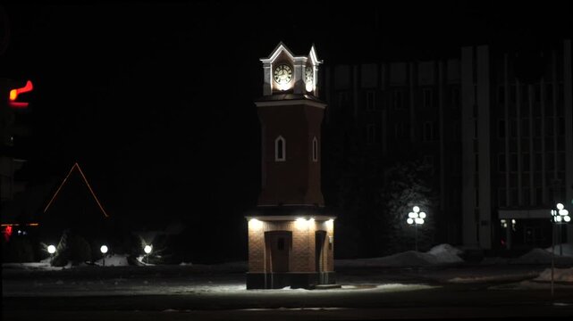 Small clock tower glowing in darkness in winter in center of tiny town.