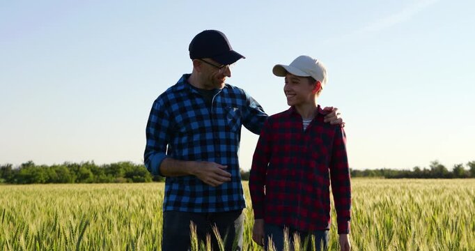 Happy father and son walking through green wheat field with arms around each other. Dad and child smile, share conversation and enjoy quality time together in countryside surrounded by nature.