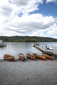 View of traditional wooden rowing boats moored on the pebble shore of Lake Windermere with a pier and forested hills under a cloudy sky Windermere, England, United Kingdom.