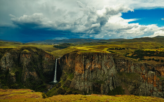 View of Maletsunyane Falls plunging into a deep gorge surrounded by rugged cliffs and rolling green hills under a dramatic cloudy sky in Maseru, Lesotho.