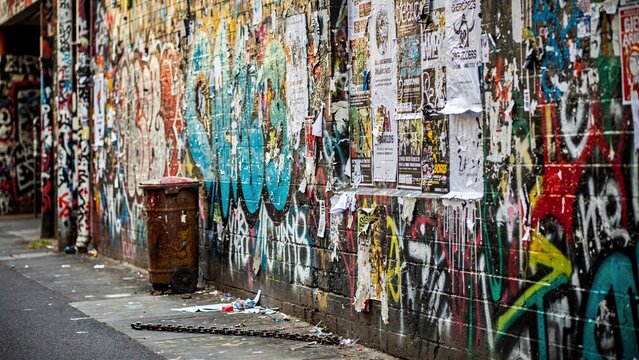 Urban street art graffiti wall covered with vibrant spray paint, torn posters, and distressed textures, showcasing a gritty city alleyway with a rusty trash can and metal chain on the dirty sidewalk.