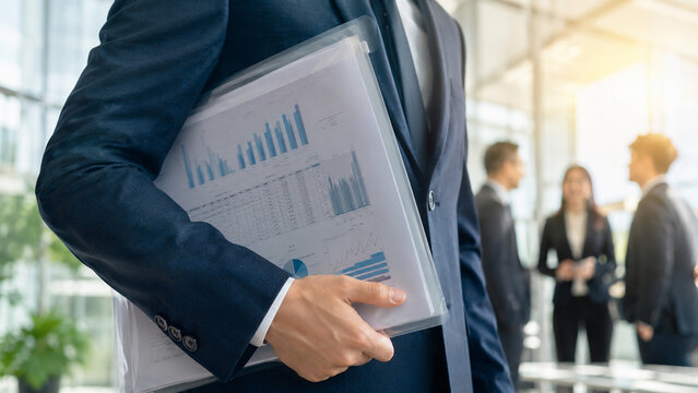 Close-up of a businessman in a navy suit holding a transparent folder with visible reports and charts, modern office background with confident colleagues, warm sunlight, cinematic lighting, stock phot