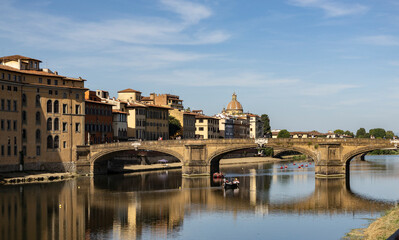 Obraz premium Florence, Italy, September 09 2025 Sunlit view of Florence’s Arno River with the elegant arched bridge, riverside Renaissance buildings and small boats reflecting in calm water