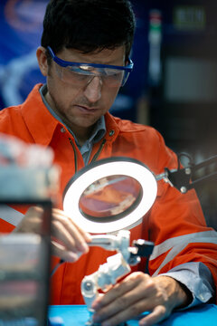 Robotics Engineer at Work: A focused engineer in safety glasses and an orange vest meticulously assembles a miniature robotic arm under the bright light of a magnifying lamp.