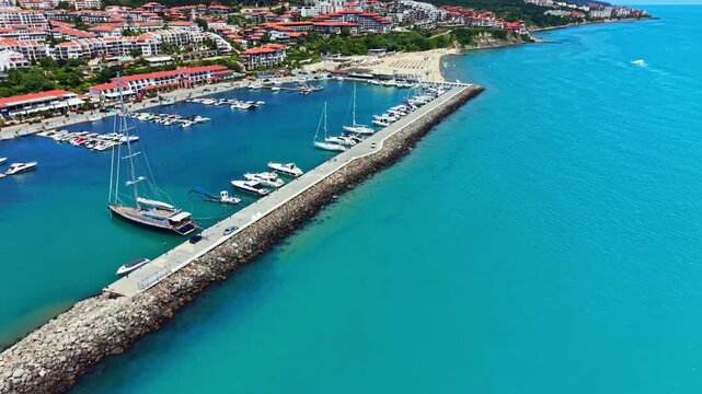 Boats are lined up at a marina by the sea. A coastal town with buildings sits on a hill. The sky is clear and the water is bright blue. People can be seen enjoying their time by the shore.