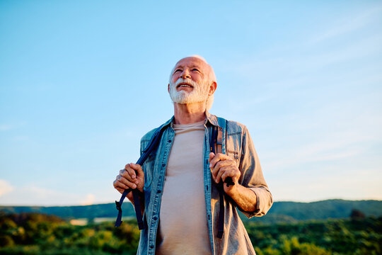 Happy senior man smiling while holding backpack straps, enjoying active retirement outdoors under clear blue sky
