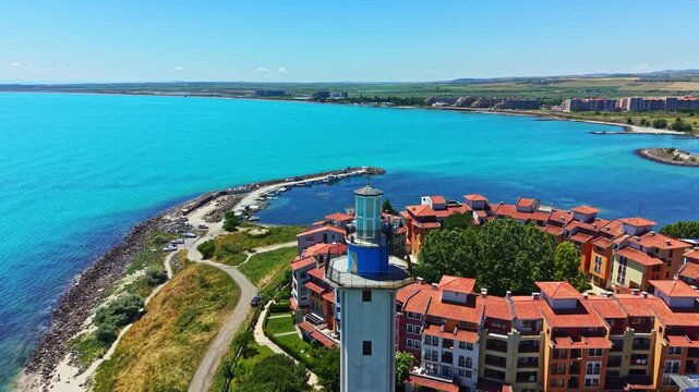 The lighthouse is situated by a cluster of homes near a bright blue sea. Waves lap against the shore, and buildings stretch into the distance under a clear sky.