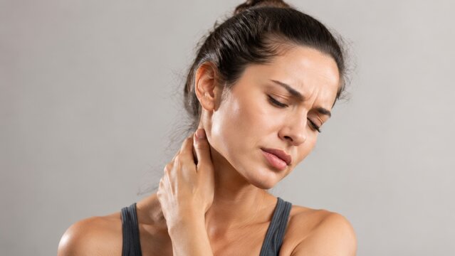 Close-up portrait of a young woman in a sleeveless top holding the side of her neck with one hand and wincing in discomfort. Neutral gray background and soft