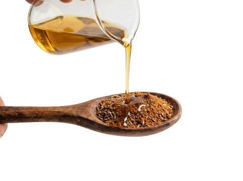 Golden liquid oil pours from a clear glass beaker onto a rustic wooden spoon filled with aromatic cumin seeds and ground brown spices against a clean white studio background for culinary photography p
