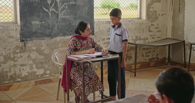 Indian young adult female sitting on chair do school work check study book exam notes indoor class room. Teen boy child wear uniform look tutor talk speak explain lesson at rural area village place