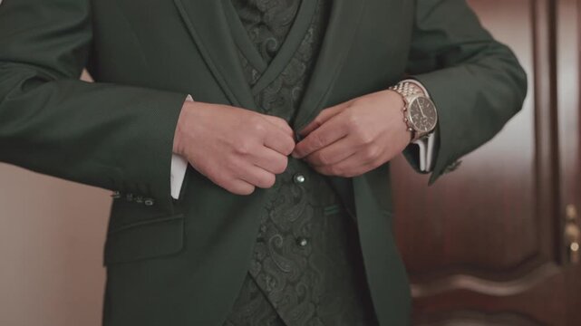 Man in dark green suit fastening paisley waistcoat buttons wearing luxury chronograph watch