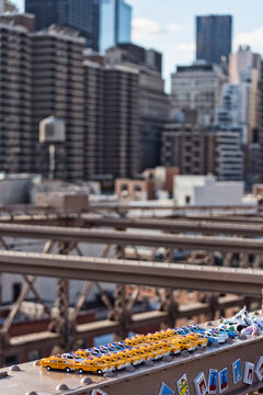 Miniature toy taxis and police cars lined up on the Brooklyn Bridge beam with a blurred Manhattan skyline, capturing urban travel and tourism in New York City, USA.