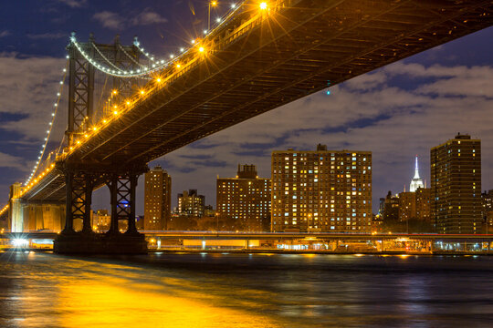Manhattan Bridge glowing at night over the East River with the NYC skyline, representing an iconic travel destination and tourism landscape in New York City, USA.