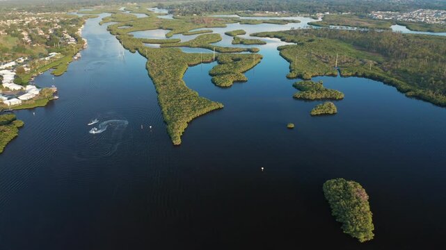 Mangrove-covered islands sit in calm Braden River water as a small motorboat cuts curved wakes near nearby suburban homes.