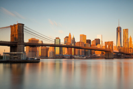 Brooklyn Bridge spans the East River before the Lower Manhattan skyline, highlighting urban tourism and global travel exploration during sunset in New York City.