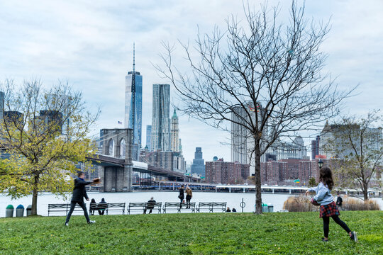 People playing catch on a green park lawn, embodying an active outdoor lifestyle with the Brooklyn Bridge and Lower Manhattan skyline background, New York City, USA.