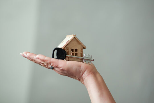 A woman&rsquo;s hand on a pastel gray studio background holds a wooden house model and a large key. Minimal still life symbolizes home purchase, key handover, rent or mortgage. Soft lighting evokes a calm