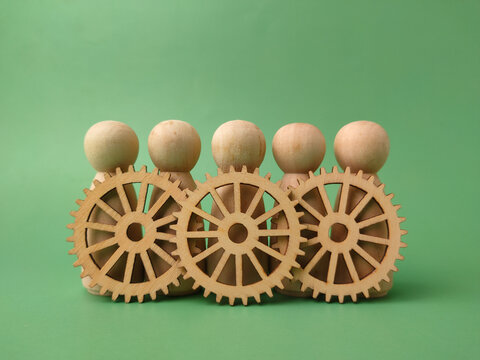 Five blank wooden peg dolls stand lined up behind three interconnected decorative wooden cogs on green background