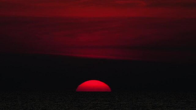 Large red sun sinking below the ocean horizon under a dark crimson sky at dusk. Extreme close-up telephoto shot.