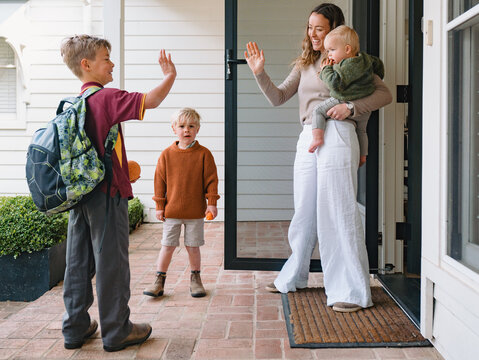 Aussie mum carrying toddler boy waving goodbye to older son leaving for school at the porch