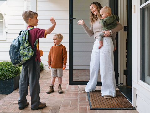 Aussie mum carrying toddler boy waving goodbye to older son leaving for school at the porch