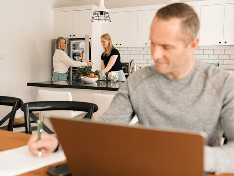 Teenage daughter helping mum with groceries in the kitchen while dad works on computer