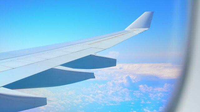 View from an airplane window showing the aircraft wing soaring above bright cumulus clouds under a clear blue sky, symbolizing freedom, global travel, exploration, aviation, limitless possibilities.
