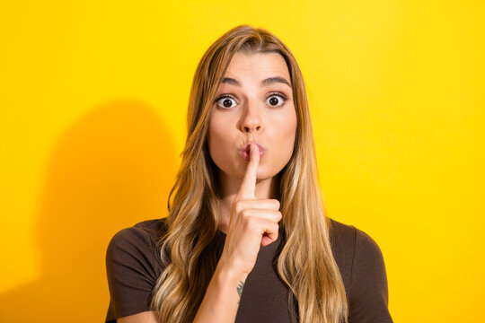 Young woman making shushing gesture with finger to lips against bright yellow background, portrait conveying secrecy, silence, surprise and playful hush