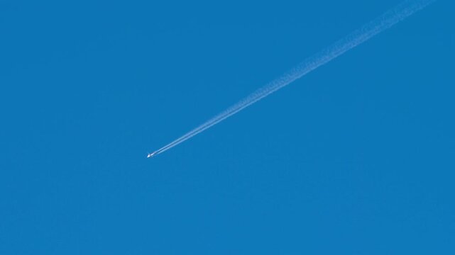 Commercial airplane flying at high altitude, leaving a long white condensation trail in a clear blue sky during a sunny day, shot with a telephoto lens