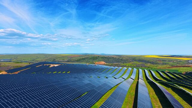 Drone view of extensive solar panel array on rolling hills. Wide aerial view of rows of photovoltaic panels contouring a green hillside with excavators working on the ridge.