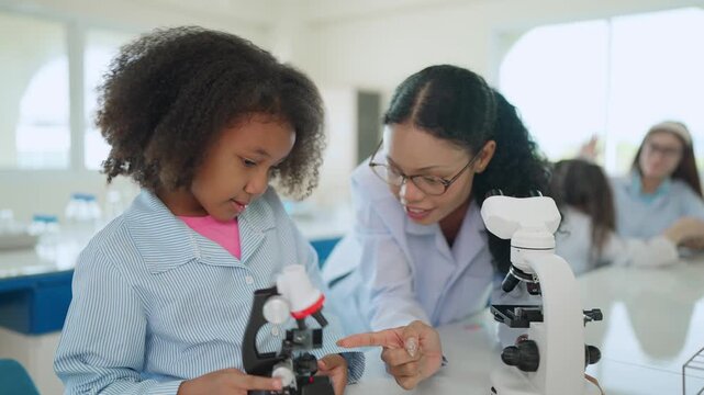 Young Scientists in the Lab: A young girl, wearing a lab coat, looks intently at a microscope while a teacher guides her, fostering a passion for science and discovery.