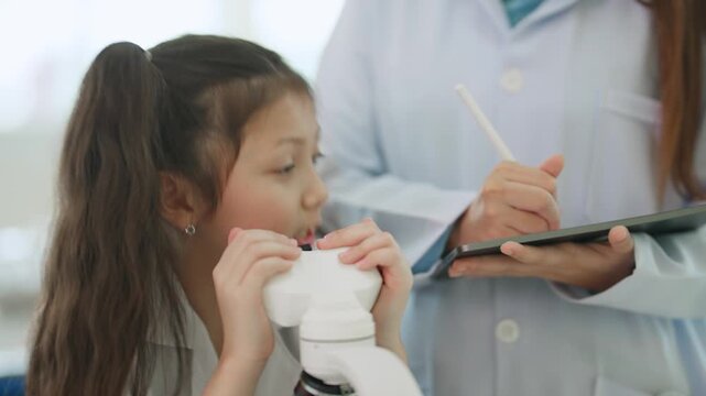 Young Scientist's Discovery: A curious young girl in a lab coat examines a microscope, her teacher observing and documenting the exciting experiment.