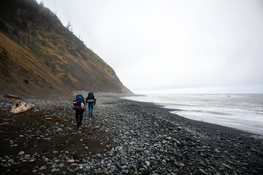 View of two hikers with backpacks walking along the pebbly shore of the Lost Coast beneath steep cliffs in Humboldt County, California, United States.