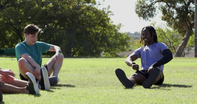 Diverse male team fist bumping, sitting on grass by oak trees stretching hamstrings for warm up