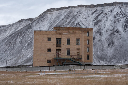 Desolate Soviet-era abandoned brick building with broken windows and an external metal staircase, set against a backdrop of massive snow-covered Arctic mountains under a gloomy sky.