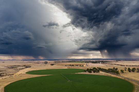 Aerial view of rain falling from dramatic storm clouds over green circular crops in dry farmland