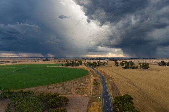 Aerial view of rain falling from dramatic storm clouds over green circular crops in dry farmland