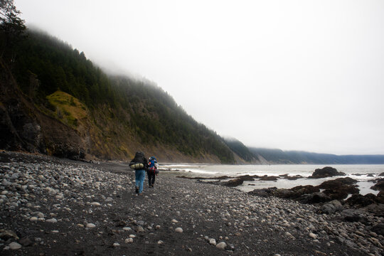 View of two hikers with backpacks walking along the rocky shoreline of the Lost Coast by the Pacific Ocean under foggy forested hills in Humboldt County, California, United States.