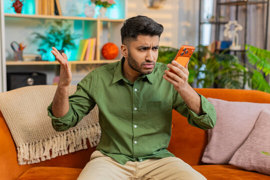 Indian man on home sofa speaks on smartphone speaker call with stop gesture and upset look in tense argument. Arabian guy on home sofa reacts with stress and anger in loud phone dispute indoors alone